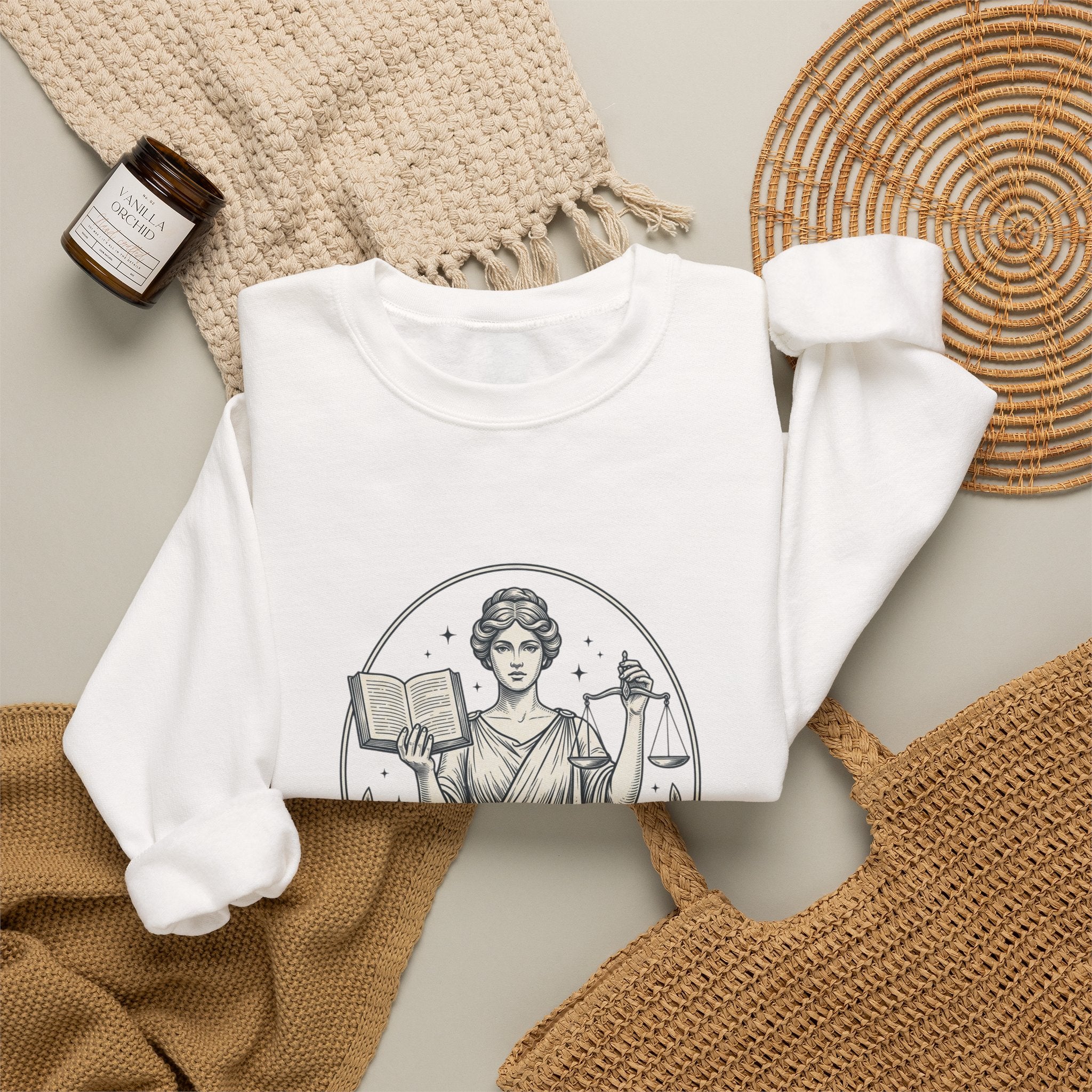 A white crewneck sweatshirt with a black line art graphic of a classical woman holding an open book and a scale, styled in a flat lay with a woven blanket, a small candle, and a woven mat.